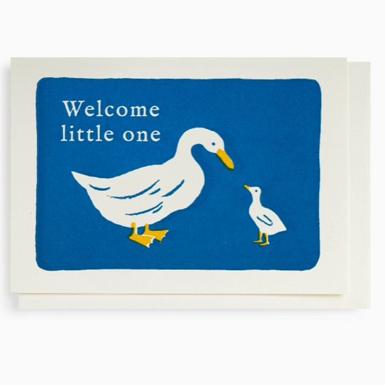 A square greeting card with a white envelope behind it, featuring a blue panel with a white duck, a smaller white duckling, and the words 'Welcome little one' in white text.