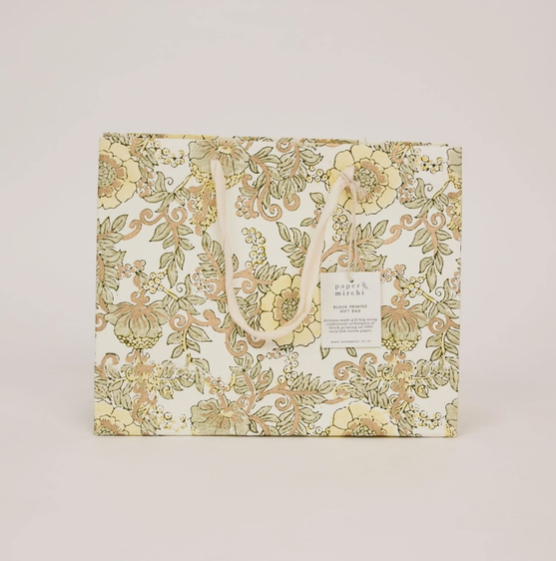 A rectangular gift bag with a cream background and an ornate floral pattern in muted yellow, beige, and olive green. It features two cream rope handles and a small white paper tag attached near the top right.
