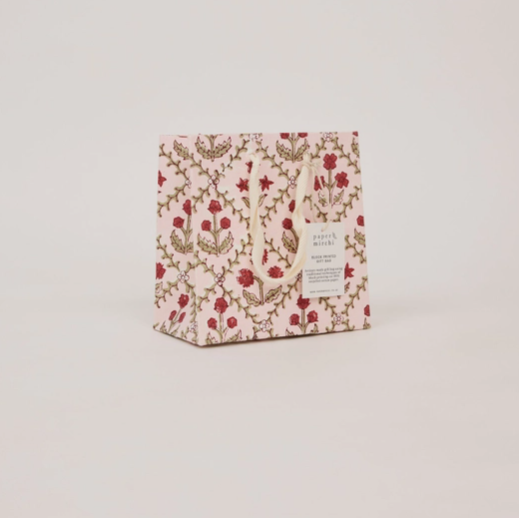 A small gift bag or packaging box with a light pink background and an allover floral pattern in red and gold, featuring a small white label on the front, set against a plain light gray background.