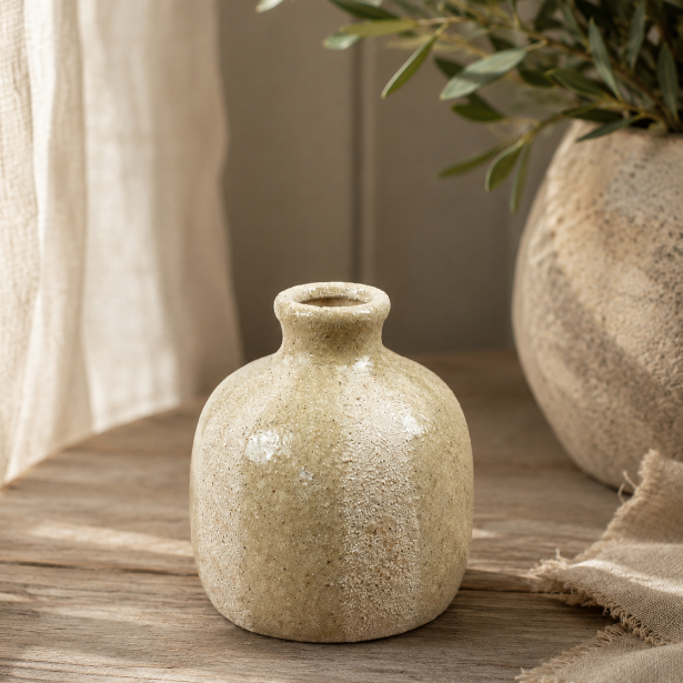 A small round ceramic vase with a short neck, featuring a textured, speckled beige and cream glaze, displayed on a rustic wooden surface next to a larger matching vase with greenery.