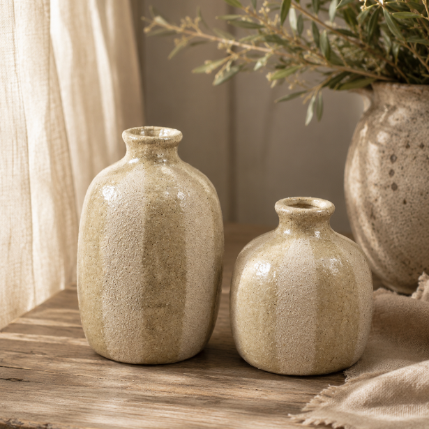 Two small round ceramic vases with a textured, speckled beige glaze sit on a rustic wooden table next to a larger vase filled with dried greenery.