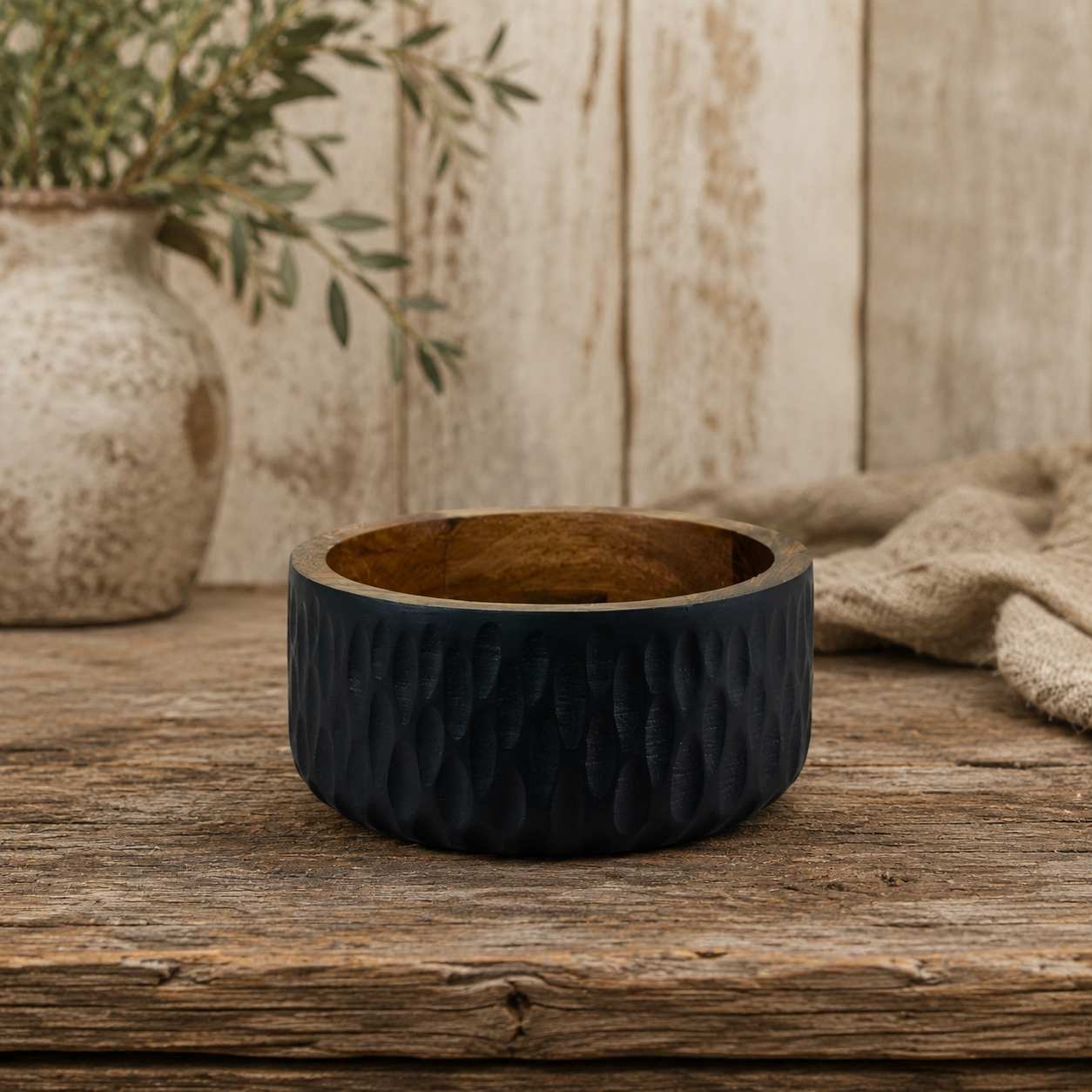 A dark navy blue textured bowl with a natural wood interior, displayed on a rustic wooden table with a neutral background and a vase of greenery.