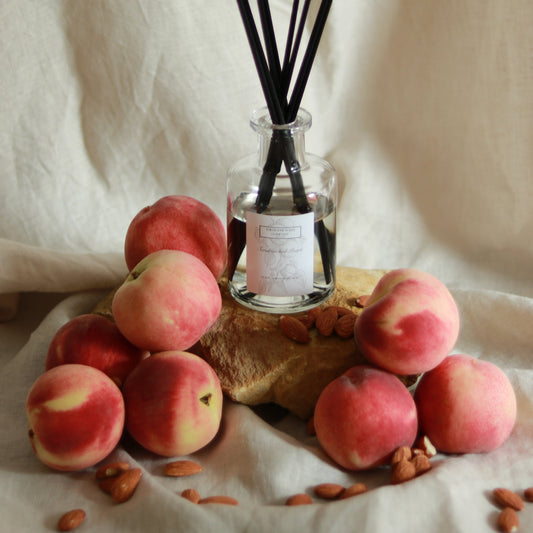 Sundrenched Peach 200ml reed diffuser by The Rustic Barn Company with black fibre reeds, displayed on a wooden stand surrounded by fresh peaches and almonds on a linen cloth