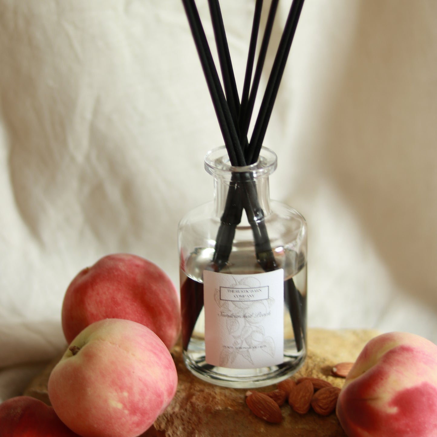 Sundrenched Peach 200ml reed diffuser by The Rustic Barn Company with black fibre reeds, styled with fresh peaches and almonds on a linen background