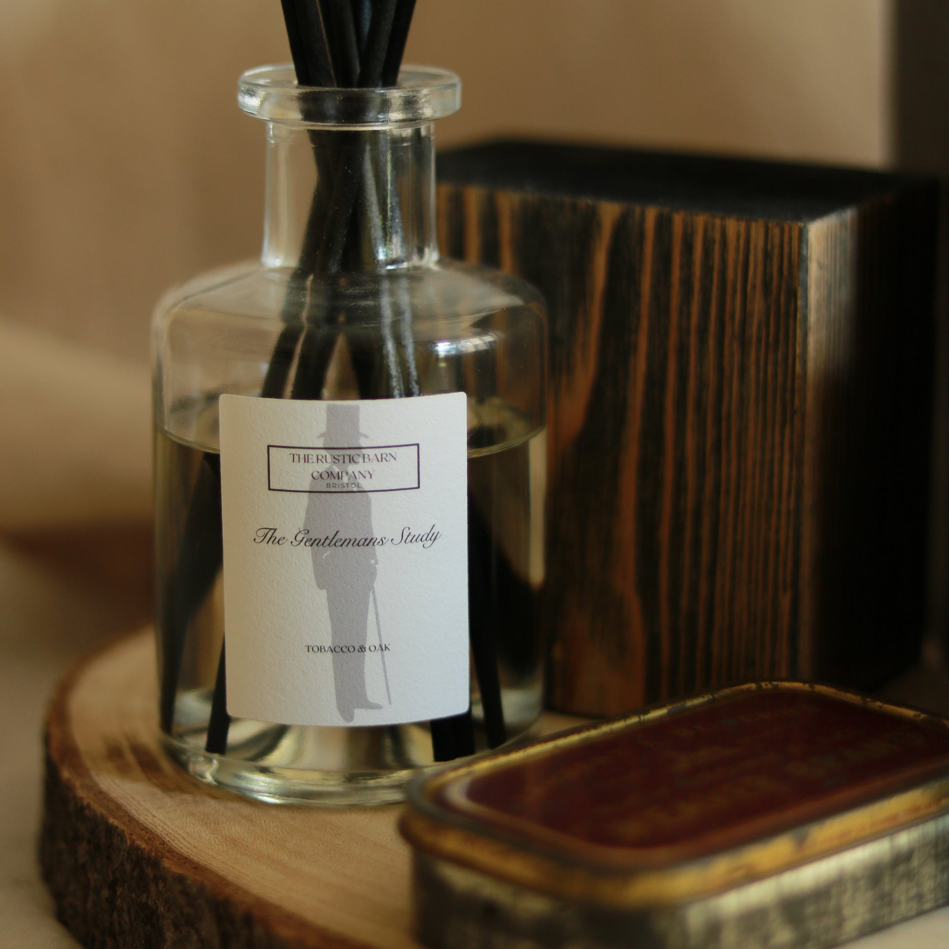 The Gentleman's Study 200ml reed diffuser by The Rustic Barn Company with black fibre reeds, displayed on a wooden slice alongside a dark wood box and vintage tin