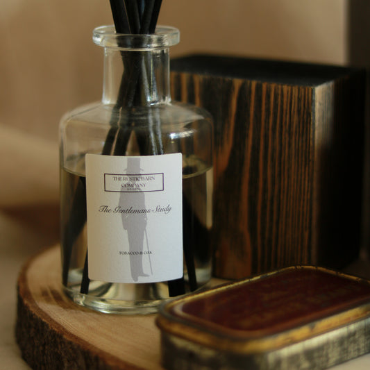 The Gentleman's Study 200ml reed diffuser by The Rustic Barn Company with black fibre reeds, displayed on a wooden slice alongside a dark wood box and vintage tin