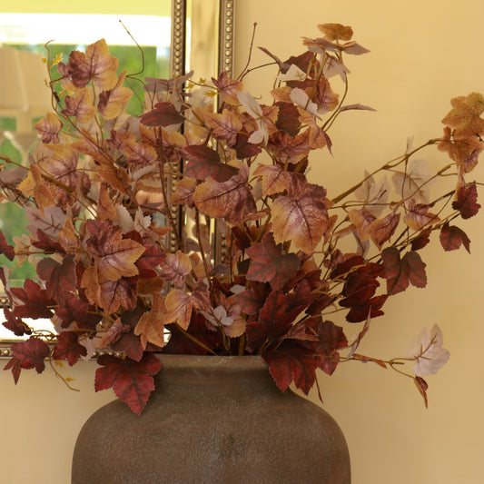 Artificial autumn grapevine branches with burgundy, brown, and tan leaves arranged in a dark gray ceramic vase against a beige wall.