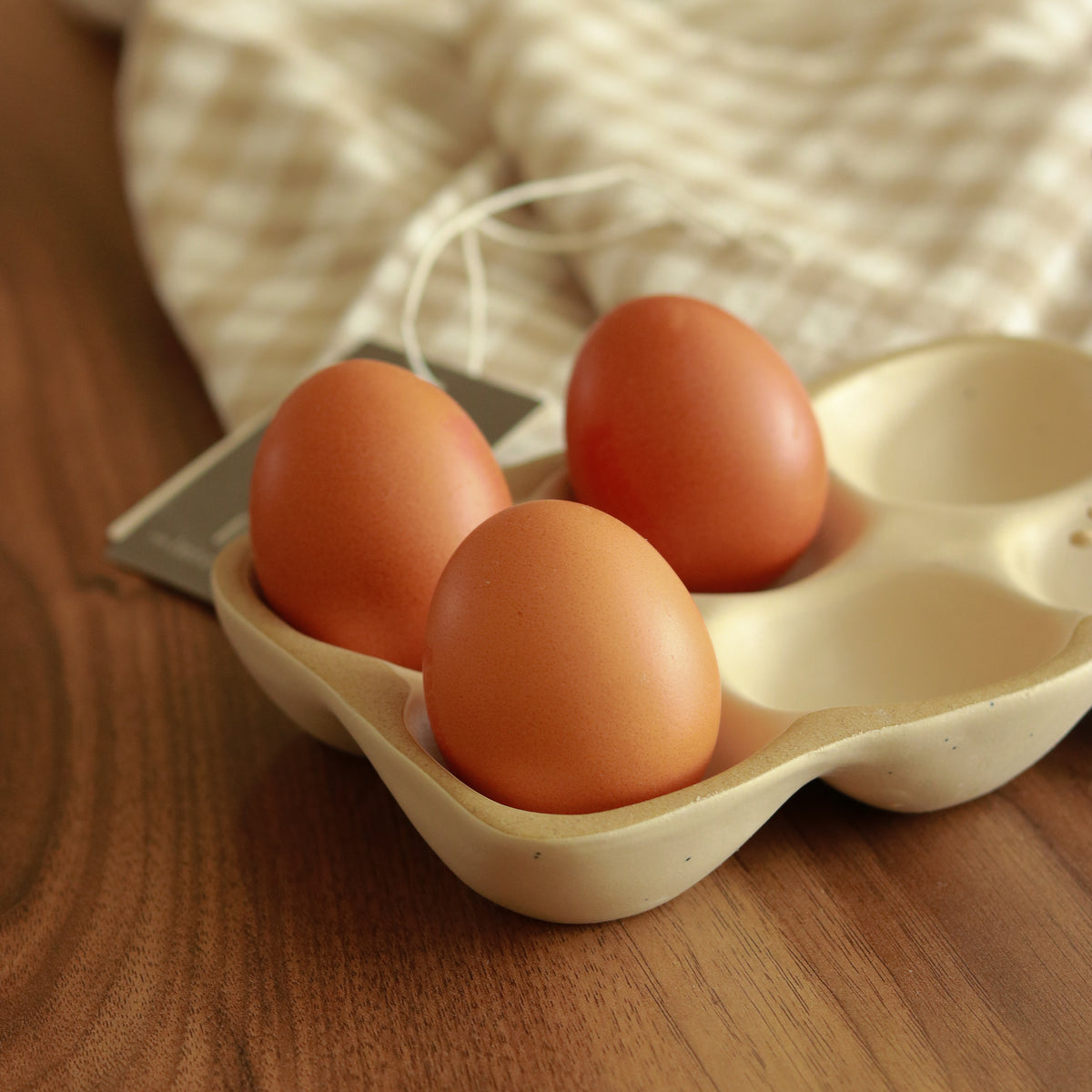 Three brown eggs resting in a beige ceramic egg carton on a wooden table, with a soft cream checkered cloth blurred in the background.
