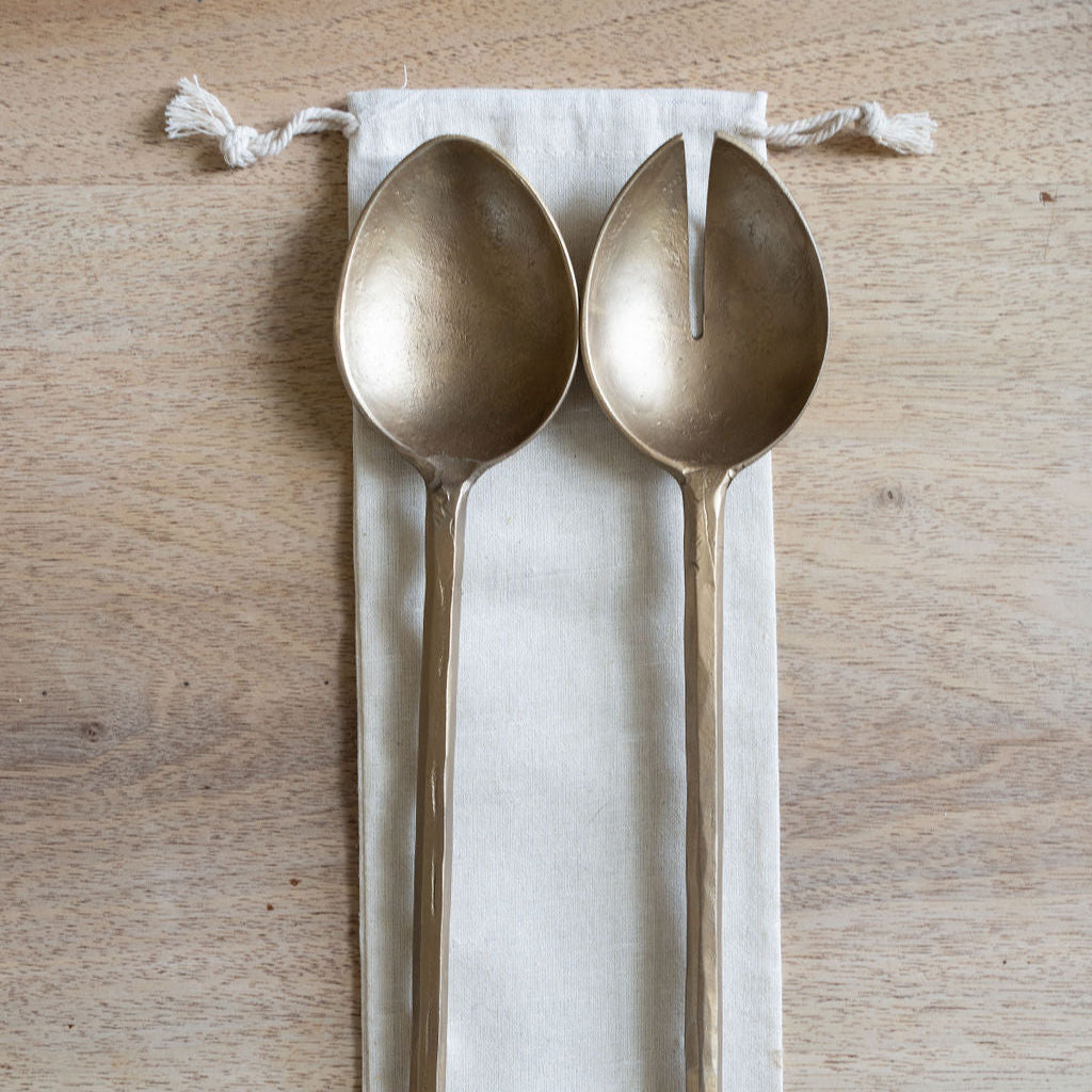 Two gold-toned metal serving utensils, one spoon and one split serving spoon, resting on a white fabric drawstring pouch with a 'Thoughtfully Handcrafted' logo, set on a light wood background.