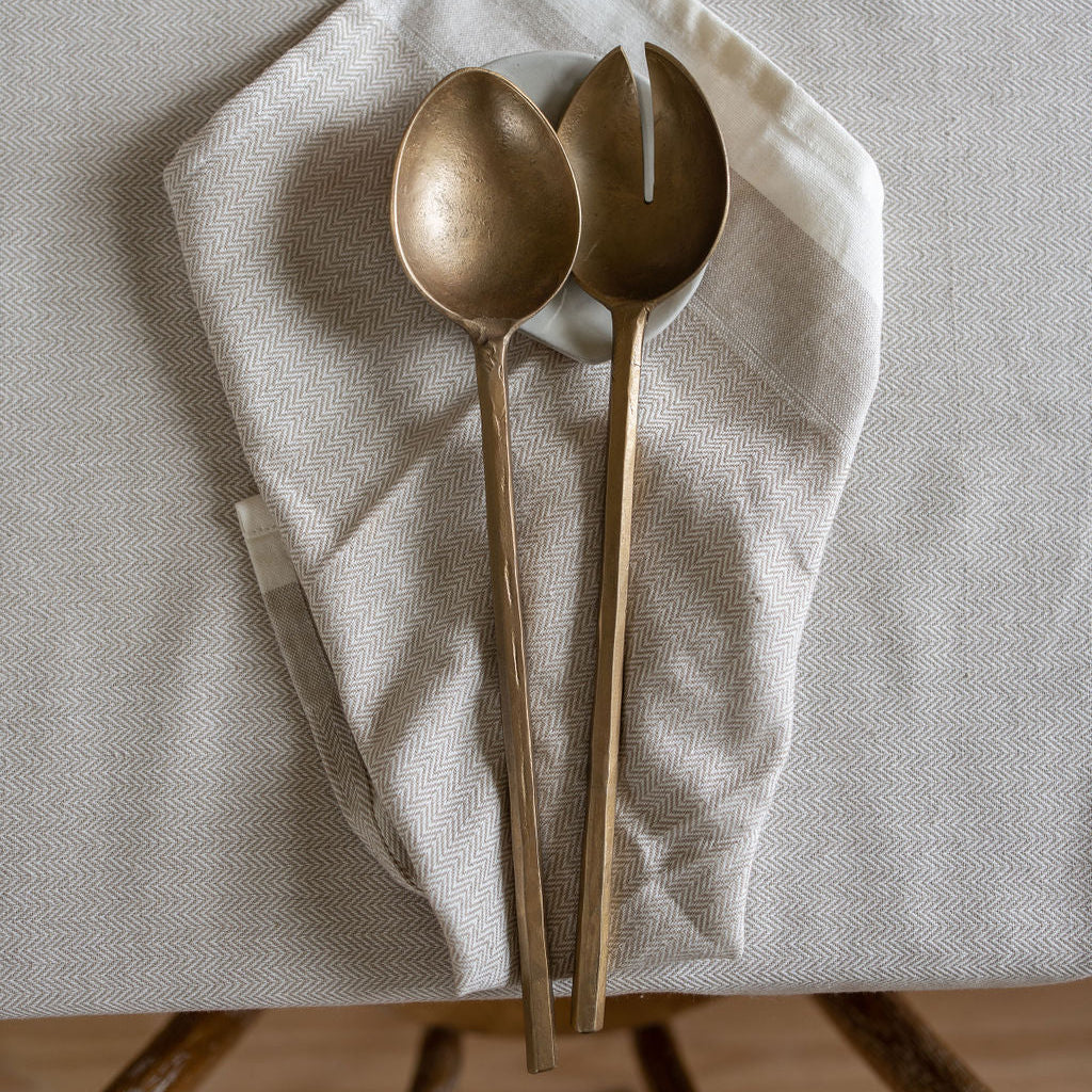 Two brass-toned tableware utensils, a spoon and a serving spoon, resting on a folded white linen napkin over a light beige tablecloth.
