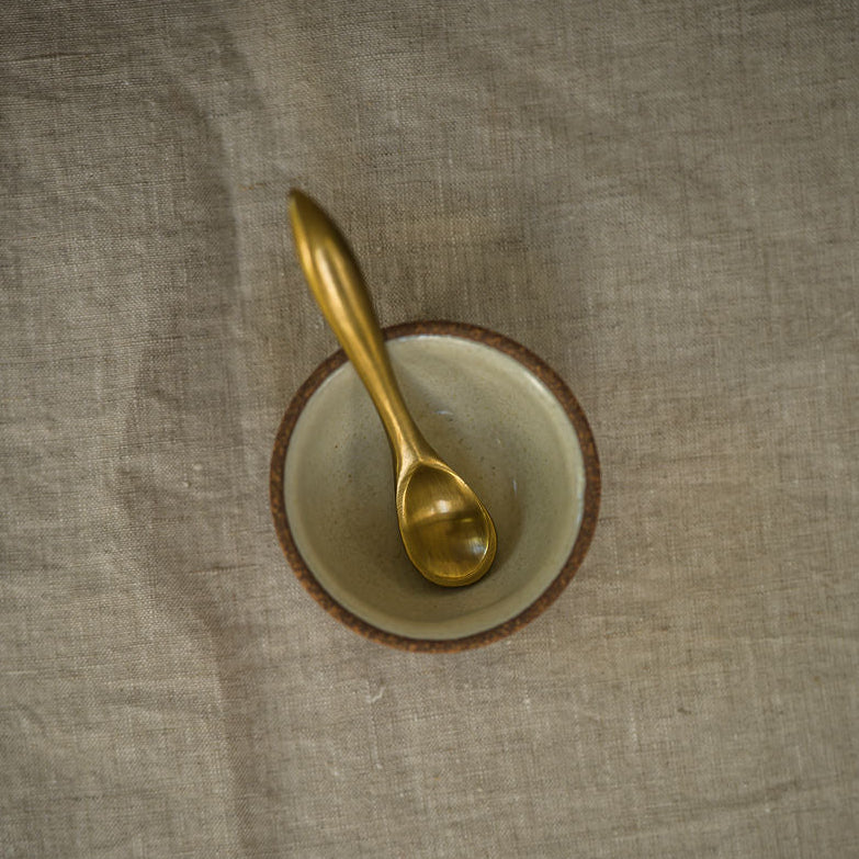 A small gold spoon resting inside a shallow round bowl with a brown rim, placed on a textured beige fabric surface.