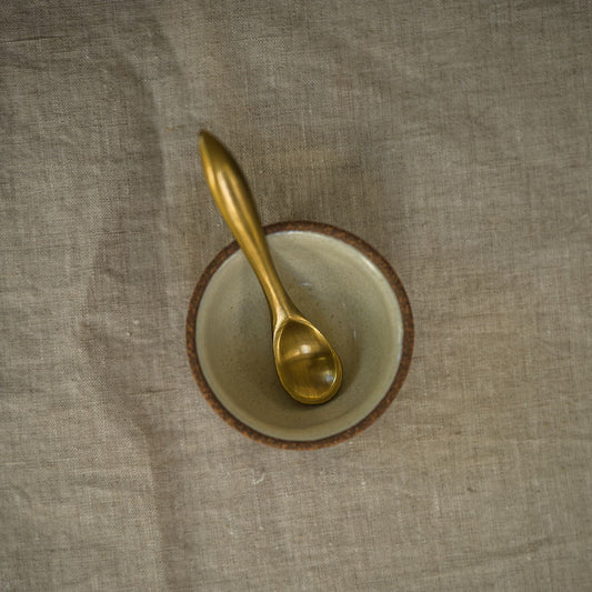 A small gold spoon resting inside a shallow round bowl with a brown rim, placed on a textured beige fabric surface.