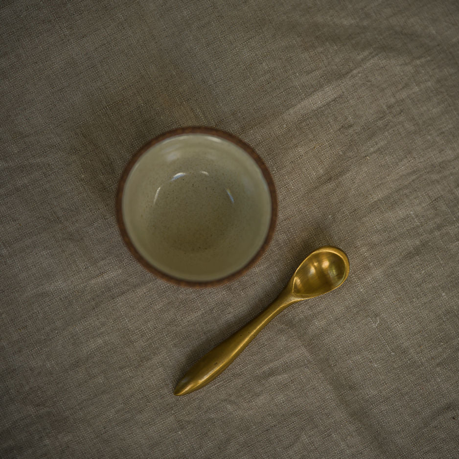 A small ceramic bowl with a brown rim and a gold spoon placed on a textured gray fabric background.