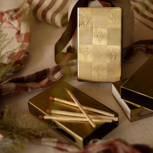 Gold embossed floral matchbox sleeve shown upright with open matchbox and red-tipped matches scattered on a festive styled surface with ribbon and foliage