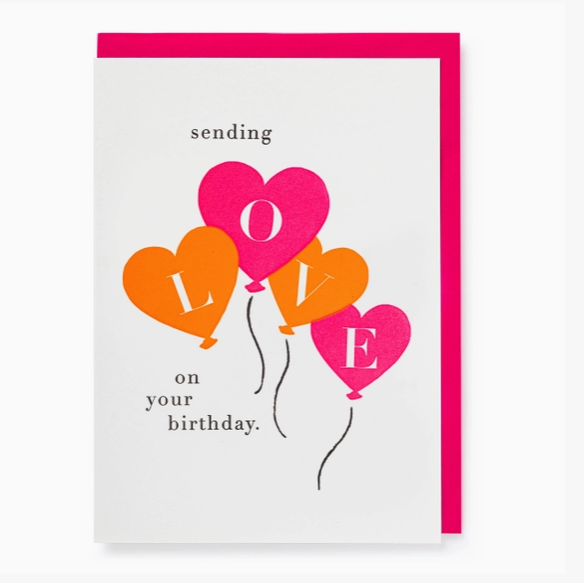 A square birthday greeting card with a bright pink envelope behind it. The card has a white background and features four heart-shaped balloons in pink, orange, and yellow, spelling out LOVE, with the words 'sending on your birthday.