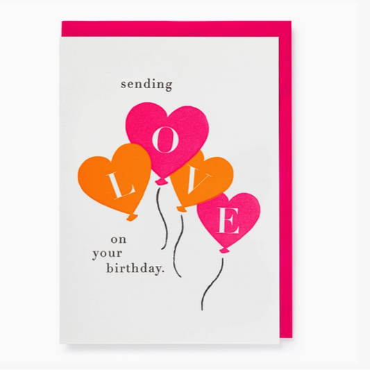 A square birthday greeting card with a bright pink envelope behind it. The card has a white background and features four heart-shaped balloons in pink, orange, and yellow, spelling out LOVE, with the words 'sending on your birthday.