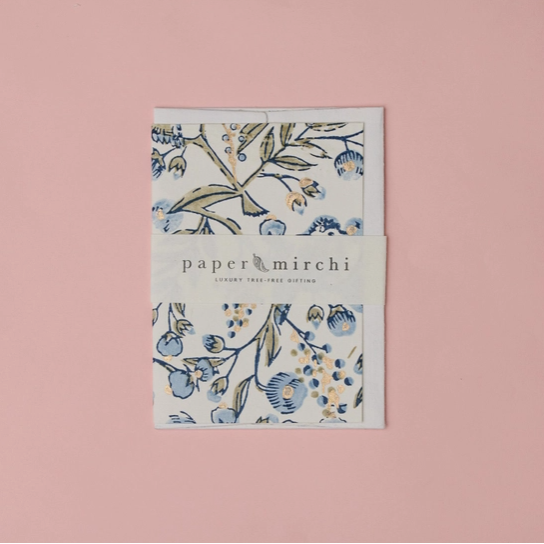 A Paper Mirchi London stationery kit featuring a floral patterned notebook or card with blue and green botanical illustrations, wrapped in a white paper band with the brand name 'paper mirchi' printed on it, set against a solid dusty pink background.
