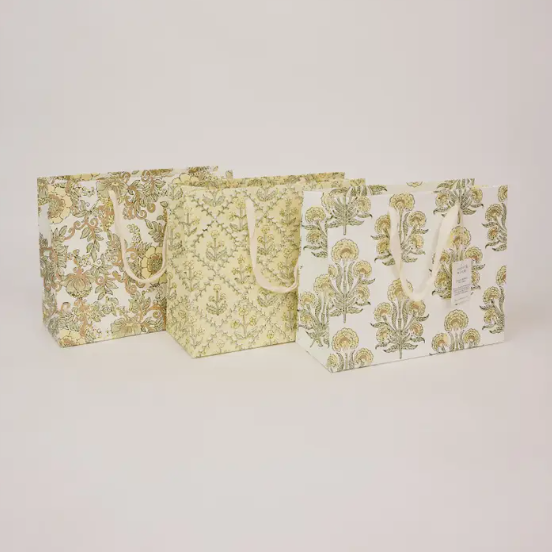 Three decorative paper shopping bags with cream handles, featuring botanical and damask patterns in muted olive green and beige, arranged against a plain light gray background.