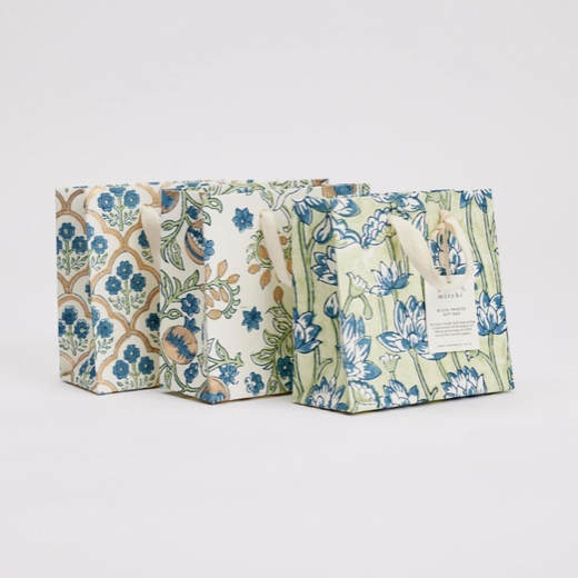 Three decorative paper gift bags with floral and geometric patterns in blue, green, beige, and brown, arranged on a light gray background.