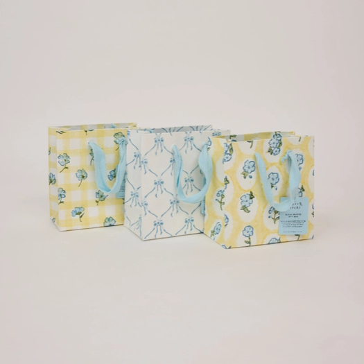Three small gift bags with light blue handles, displayed against a plain light gray background. The bags feature different pastel patterns: a yellow and white gingham on the left, a white and light blue geometric design in the center, and a yellow and white floral print on the right with a small blue label.