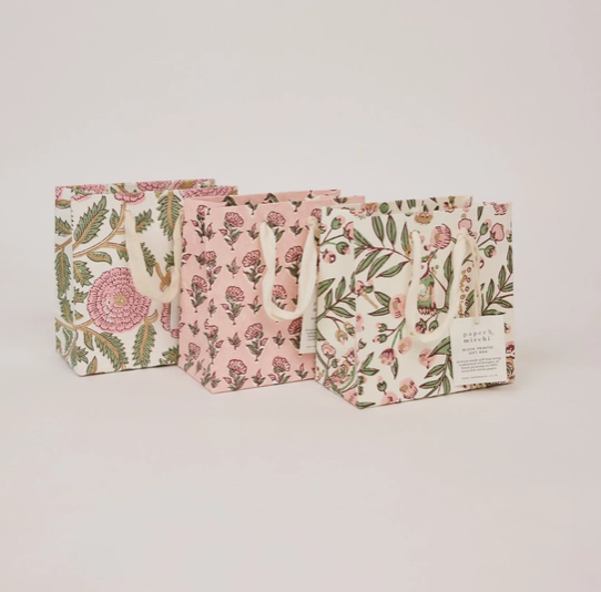 Three floral paper gift bags with white handles, arranged in a row on a light beige background. The bags feature different floral patterns in pink, green, and gold on cream and pink bases.