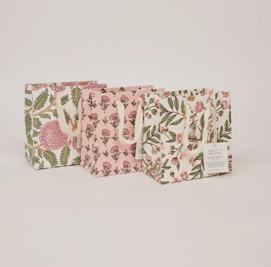Three floral paper gift bags with white handles, arranged in a row on a light beige background. The bags feature different floral patterns in pink, green, and gold on cream and pink bases.