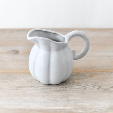 A small white ceramic pitcher with a ribbed pumpkin-like body, flared spout, and loop handle, placed on a light wood surface against a white wall.