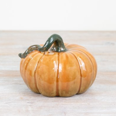 A small glazed ceramic pumpkin figurine with a dark green stem, sitting on a light-colored wooden surface against a white background.