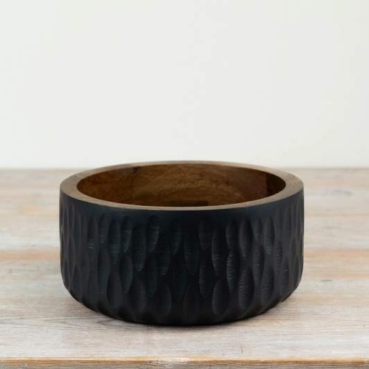 A wide, circular black wooden bowl with a textured, hammered outer surface and a smooth, warm brown wood interior, displayed on a light wooden tabletop against a neutral off-white background.