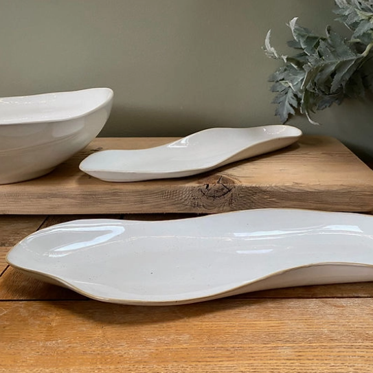 Three white ceramic wavy serving dishes of different sizes arranged on a wooden cutting board and tabletop, with a neutral wall and decorative leaves in the background.