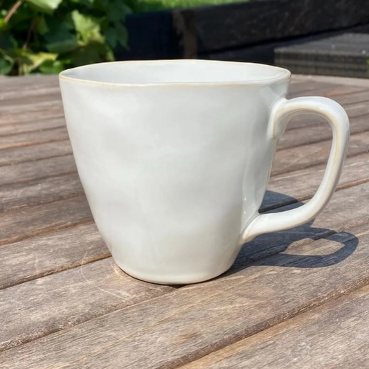 Dune oyster white stoneware mug with organic curves and reactive glaze finish, shot from the front on a weathered outdoor garden table with green foliage behind