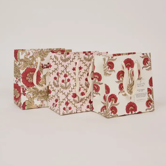 Three decorative gift bags with a vintage botanical pattern in red and gold on a cream background, arranged on a light gray studio background.