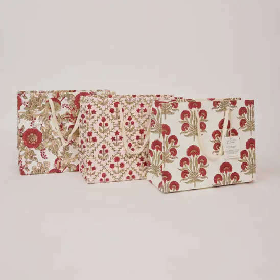 Three gift bags with red and beige botanical patterns, arranged in a row against a plain light gray background.