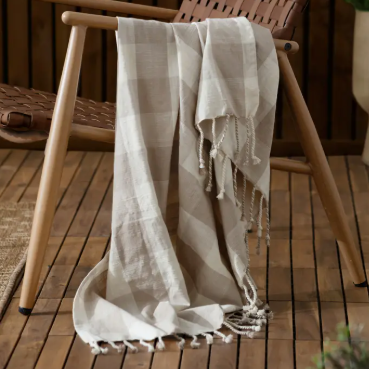 A light beige and white checkered throw blanket with fringed tassels draped over a wooden lounge chair on a wooden deck.