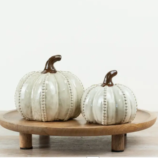 Two ceramic decorative pumpkins in a glossy off-white glaze with dark brown stems, displayed on a round wooden pedestal against a plain white background.