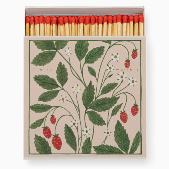 A decorative matchbox with a botanical illustration of green leaves, small white flowers, and red strawberries on a light beige background. The top of the box is open, showing a row of wooden matches with red tips.