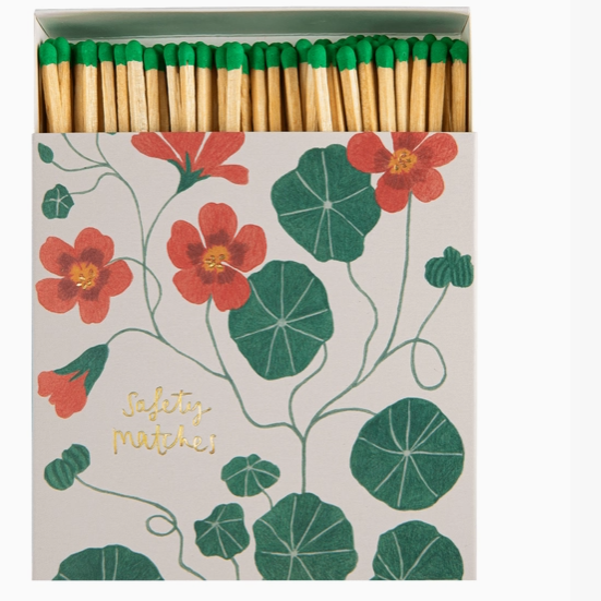 A box of safety matches with a botanical pattern of red flowers and green leaves on a light beige background. The wooden matches have bright green tips, and the box features the words 'safety matches' in gold foil script.