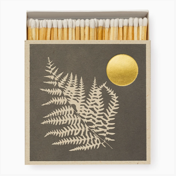 A matchbox-style container with a dark charcoal cover featuring a cream fern illustration and a gold circular sun motif, shown open with wooden matches and white tips inside.