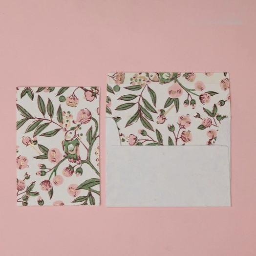 A floral-patterned envelope with a matching card placed beside it on a solid pink background. The pattern features pink blossoms, green leaves, and small bird illustrations on a white base.