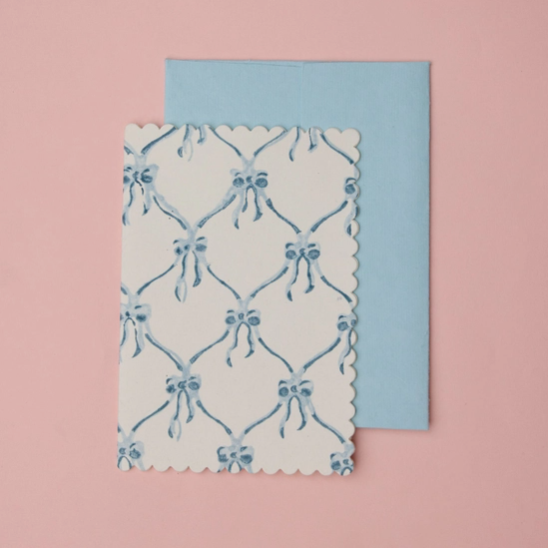 A white card with a blue bow and lattice pattern and scalloped edges, placed in front of a light blue envelope on a solid pink background.