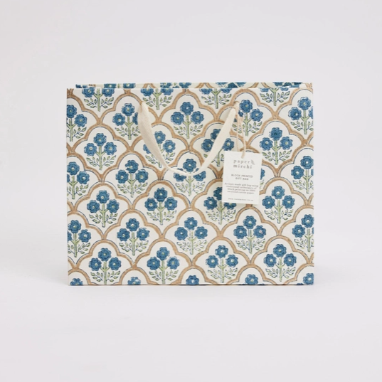 A decorative gift bag with a repeating blue floral and tan arch pattern, featuring two paper handles and a small hanging tag, photographed against a plain light gray background.