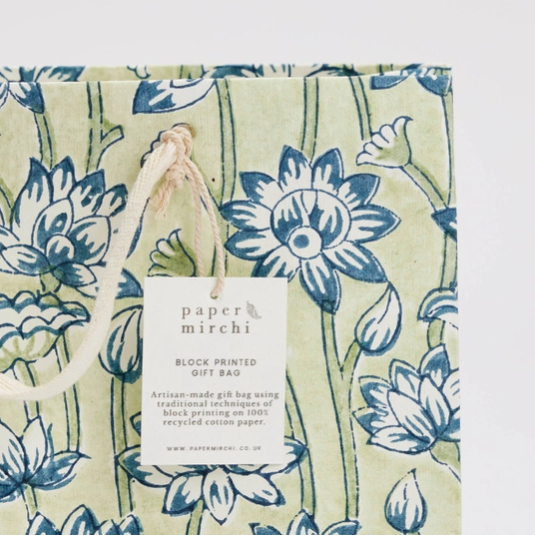 A close-up of a gift bag with a blue and green floral block-print pattern on a light beige background, featuring a white paper hangtag that reads 'paper mirchi'.