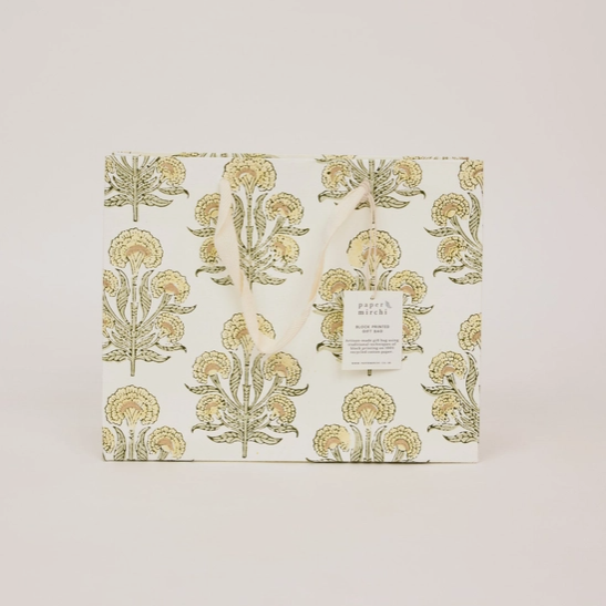 A square gift box with a white background and a repeating botanical pattern in muted olive green and warm gold. A small white paper tag hangs from a loop on the front, and the box is photographed against a plain light beige studio background.