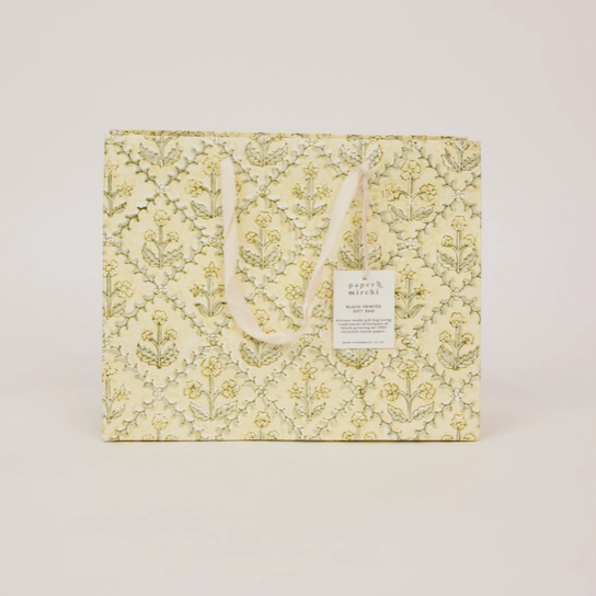 A square gift bag in a pale yellow and green floral damask patterned paper, featuring cream-colored rope handles and a small white paper tag 
