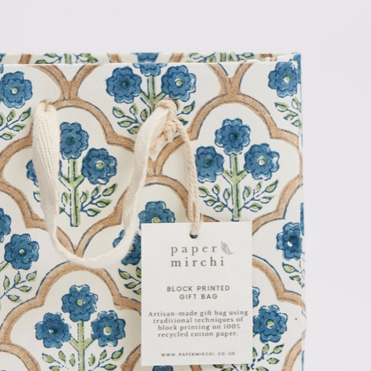 A Paper Mirchi block-printed gift bag featuring a traditional floral pattern in blue, green, and brown on a white background, with a white paper tag attached.