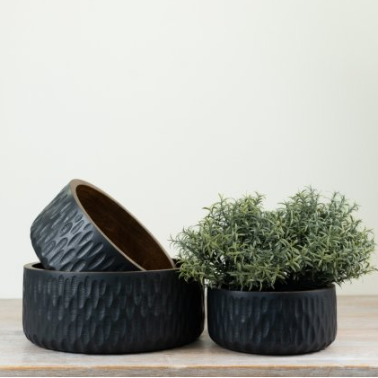 Three round black textured ceramic planters on a light wood surface against a plain off-white wall, with one planter stacked on top of another and a small green plant placed in the front right planter.