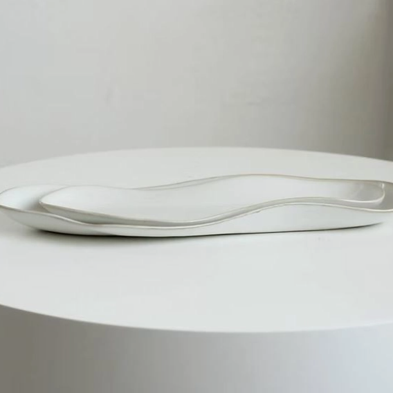 Two wavy, elongated ceramic plates in white with a subtle metallic gold rim, displayed on a round white pedestal against a neutral light gray background.