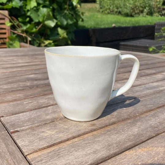 A light gray ceramic mug with a small gold rim detail, sitting on a weathered wooden outdoor table with a blurred garden background.