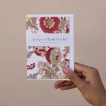 A hand holding a small square paper notebook or card set with a floral pattern cover and a white band reading 'paper mirchi' against a plain beige background.