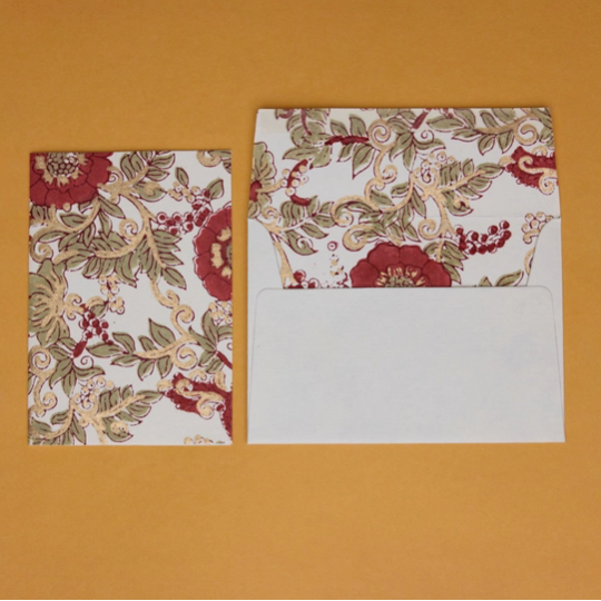 A floral patterned envelope and matching card set on a solid mustard yellow background. The envelope features a white base with a detailed botanical print in red, beige, and olive green, with the flap open to reveal a blank white interior.